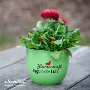 Puede incluir: Una maceta de cer&aacute;mica verde con una mariposa roja y el texto "Blumenduft liegt in der Luft" en la parte delantera. La maceta est&aacute; llena de una peque&ntilde;a planta verde con flores rojas.