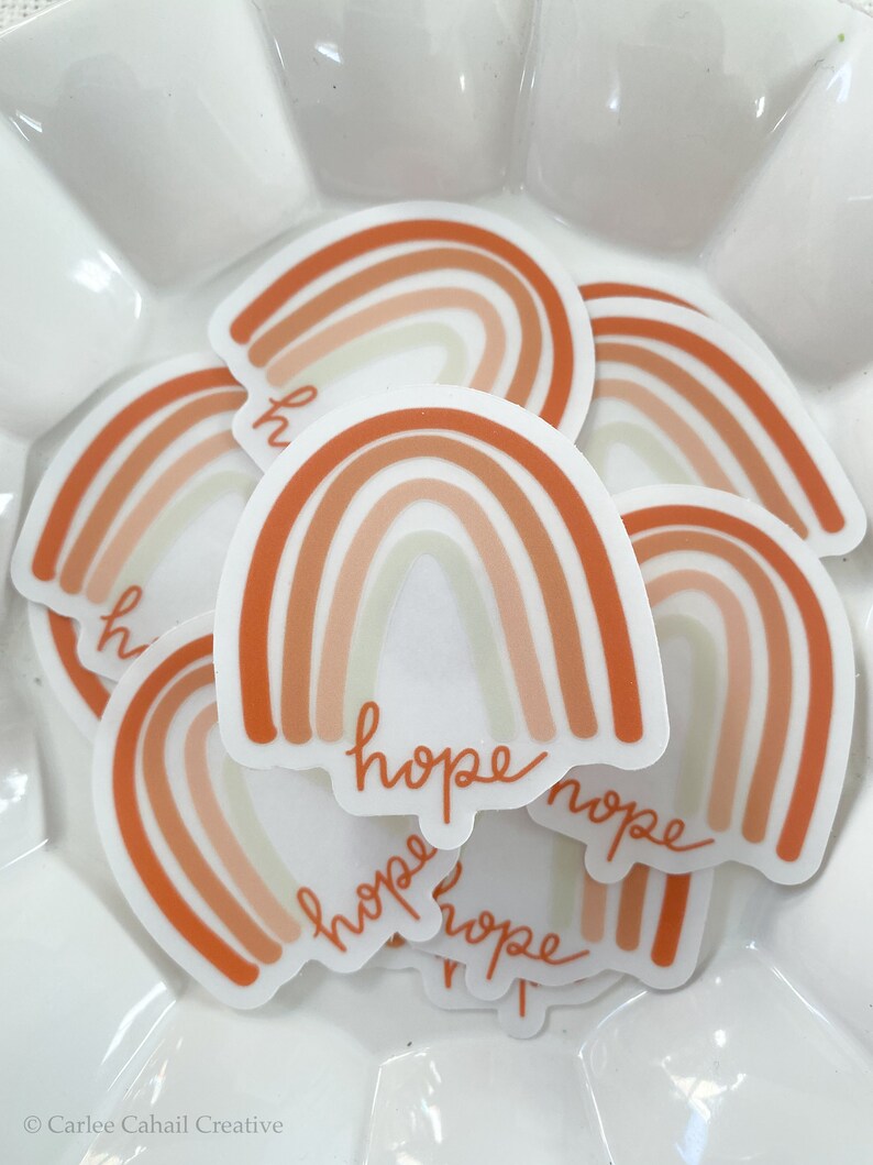 May include: A set of five rainbow stickers with the word "hope" written on each one. The rainbows are in shades of orange, peach, and cream.