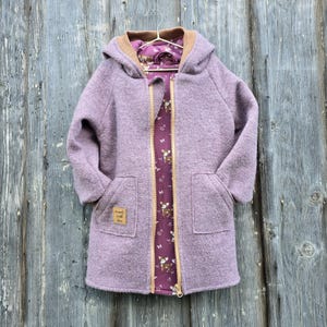 May include: A lavender wool coat with a hood and two front pockets. The coat has a brown collar and a patterned interior with deer and floral designs. A small tag on the pocket reads "made with love."
