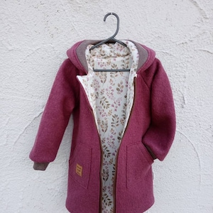 Può includere: Una giacca con cappuccio in misto lana magenta con cerniera marrone e polsini a coste. La fodera interna presenta un tessuto bianco con una stampa botanica di piccole foglie e fiori. Una piccola etichetta marrone sulla tasca riporta Made with love.