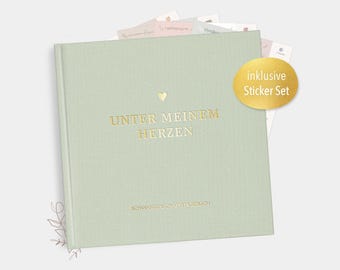May include: A light green journal with gold lettering that reads "UNTER MEINEM HERZEN" and a small gold heart. The journal includes a sticker set and has a textured cover. Several small cards peek out from the top.