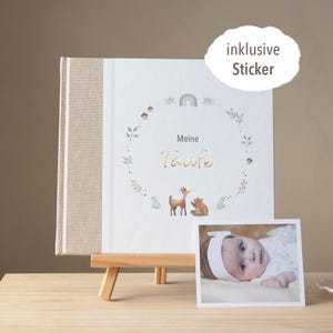 Könnte beinhalten: Ein weißes Fotoalbum mit einem Leinenbezug und einer goldenen Aufschrift "Meine Taufe". Das Album wird auf einem Holzständer präsentiert. Neben dem Album ist ein kleines Foto eines Babys zu sehen. Das Album ist mit einem Kranz aus Blättern und einer Illustration von Hirsch und Bär verziert.