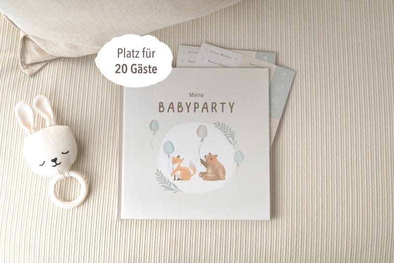 May include: A light blue and white book with the text "Meine Babyparty" and a picture of a fox and a bear with balloons. The book is on a beige surface with a white bunny rattle.