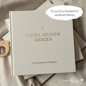 Pu&ograve; includere: Un diario beige chiaro con scritte dorate e un piccolo cuore. Il testo sulla copertina recita "UNTER MEINEM HERZEN" e "SCHWANGERSCHAFTSTAGEBUCH". Un giocattolo di legno per bambini &egrave; visibile a sinistra.