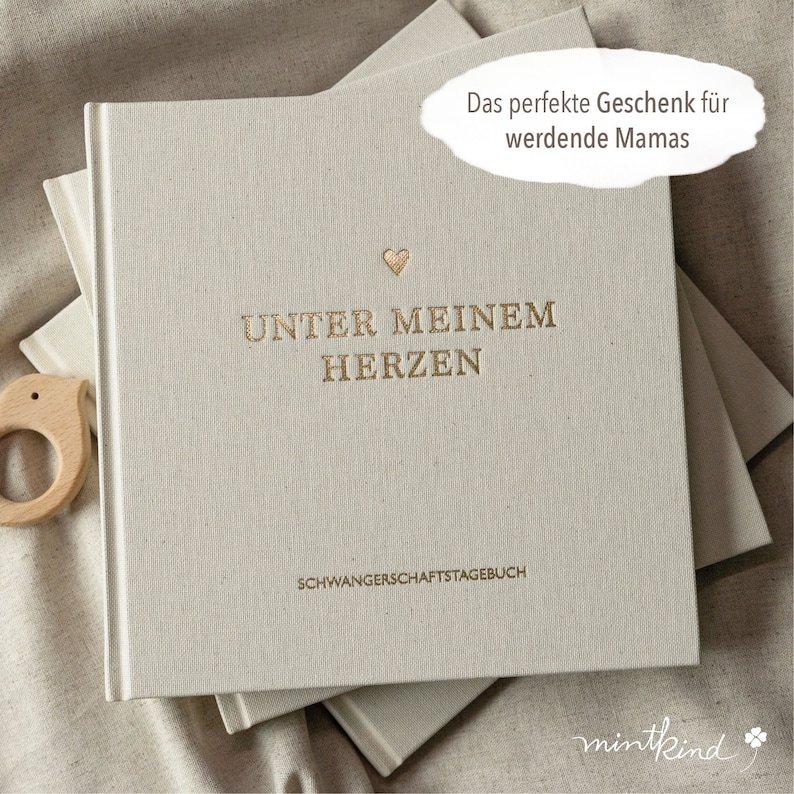 Pu&ograve; includere: Un diario beige chiaro con scritte dorate e un piccolo cuore. Il testo sulla copertina recita "UNTER MEINEM HERZEN" e "SCHWANGERSCHAFTSTAGEBUCH". Un giocattolo di legno per bambini &egrave; visibile a sinistra.