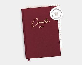 Può includere: Un'agenda con copertina rigida color bordeaux intenso con la parola Create e l'anno 2027 stampati in corsivo dorato sulla copertina. È incluso un adesivo bianco circolare con il testo DEIN PLANER 2027 INKL. STICKER SET e un disegno di una noce.