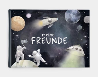 以下が含まれることがあります： 宇宙をテーマにしたイラストが描かれたハードカバーの本。表紙は、惑星、ロケット、2人の宇宙飛行士が描かれた濃い青色の背景です。「Meine FREUNDE」というドイツ語のテキストが大きく表示されています。