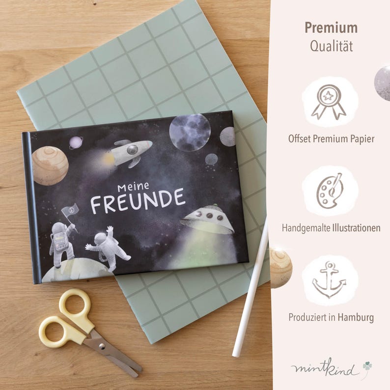 Op de afbeelding: Een kinderboek getiteld "Meine Freunde" met een ruimtethema, met illustraties van planeten, raketten en astronauten. De omslag is zwart met witte tekst en kleurrijke afbeeldingen. Een gele schaar en een groen notitieboek zijn ook zichtbaar.