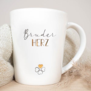 K&ouml;nnte beinhalten: Wei&szlig;e Keramiktasse mit einem schwarzen und goldenen Design. Die Tasse tr&auml;gt die Aufschrift "Bruder Herz" mit einem goldenen Herzen und vier schwarzen Herzen.