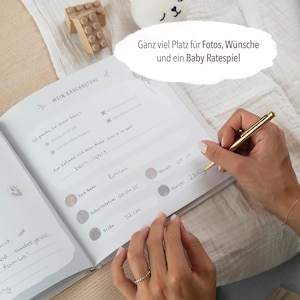 May include: A white baby journal with a gray and white floral design. The journal has a section for recording the baby's name, birthdate, weight, and length. The text "Mein Bauchgef&uuml;hl" is written in black on the cover.