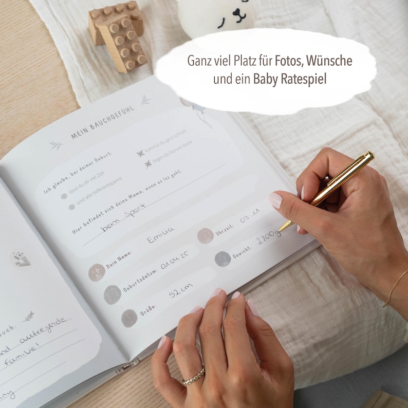 May include: A white baby journal with a gray and white floral design. The journal has a section for recording the baby's name, birthdate, weight, and length. The text "Mein Bauchgef&uuml;hl" is written in black on the cover.
