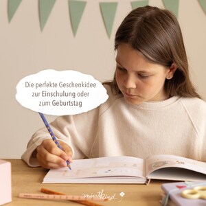 Op de afbeelding: Een persoon schrijft met een blauw potlood in een open boek. Het boek ligt op een houten oppervlak, met een potlood en een notitieboek in de buurt. Tekst in een wolk-vormige tekstballon luidt: "Het perfecte cadeau-idee voor schoolinschrijving of verjaardag."
