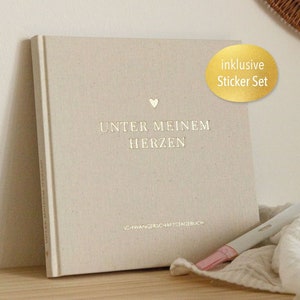 Könnte beinhalten: Beiges Notizbuch mit dem deutschen Text "UNTER MEINEM HERZEN" und einem kleinen Herzsymbol. Ein goldener Aufkleber lautet "inklusive Sticker Set". Ein Schwangerschaftstest und ein weißes Tuch sind im Vordergrund.