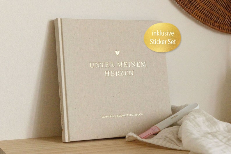 Pu&ograve; includere: Un diario beige con il testo tedesco "UNTER MEINEM HERZEN" e un piccolo simbolo a forma di cuore. Un adesivo dorato recita "inklusive Sticker Set". Un test di gravidanza e un panno bianco sono in primo piano.