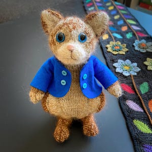 May include: A handmade, knitted cat doll with a tan and brown body, wearing a bright blue jacket with blue buttons. The cat has large blue eyes and a pink nose. The doll is standing in front of a black and gray floral scarf.
