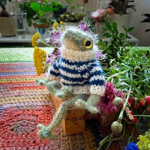 May include: A handmade, knitted frog figurine with a blue and white striped sweater and a white turtleneck. The frog has large, yellow eyes and is sitting on a small wooden block, surrounded by flowers and greenery.