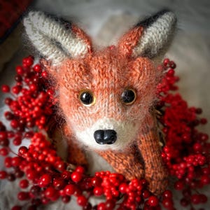 May include: A handcrafted knitted fox toy, predominantly orange with white and black accents, and large, shiny eyes. The fox is nestled within a wreath of vibrant red berries. The close-up shot emphasizes the texture of the yarn and the intricate details of the knitting.
