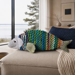 Könnte beinhalten: Ein farbenfrohes, gehäkeltes Kissen in Fischform mit einem schuppenartigen Muster in Blau-, Grün-, Orange- und Lilatönen. Der Fisch hat einen grauen Kopf und Flossen und liegt auf einem beigen Kissen. Ein dekoratives Element für das Zuhause.