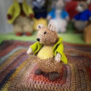 May include: A handmade knitted bear doll with a brown body, yellow shirt, and green jacket. The bear has small gray ears and a black nose. It is standing on a colorful, textured blanket. Other similar dolls are in the background.