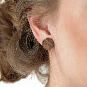 Op de afbeelding: Close-up van een oor met een ronde houten oorbel. De oorbel is donkerbruin met een houtnerfpatroon en gezet in een bronskleurig metaal. Het lichtbruine haar van de persoon is naar achteren getrokken.