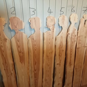 Könnte beinhalten: Acht Holzbretter mit verschiedenen Silhouetten-Schnitzereien von Personenprofilen. Die Bretter sind alle aus Naturholz in hellbrauner Farbe und haben eine raue, unfertige Textur.