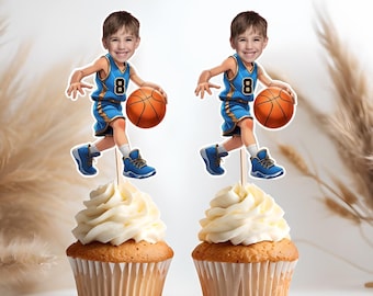 Op de afbeelding: Twee blauwe en witte cupcake-toppers met een basketbalthema met een foto van een kind in een basketbaluniform. De toppers staan op cupcakes met witte frosting.