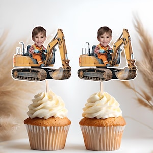 May include: Two yellow toy construction vehicles with a child's face on each. The vehicles are on cupcake toppers.