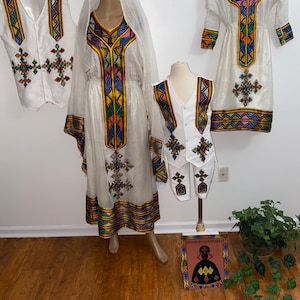Puede incluir: Un conjunto de tres prendas tradicionales etíopes blancas con bordados geométricos coloridos. Las prendas incluyen un vestido largo, un chaleco y un vestido más corto. Las prendas se muestran en maniquíes contra una pared blanca.