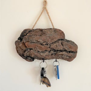 Op de afbeelding: Een rustieke houten sleutelhanger met een natuurlijke schorsafwerking. Hij heeft drie metalen haken om sleutels aan te hangen en is met een touwtje aan de muur bevestigd.