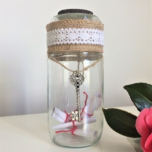 Peut inclure: Un bocal en verre avec un ruban en toile de jute et en dentelle autour du haut. Le bocal contient des morceaux de papier roulés et une clé en argent.