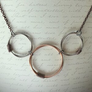 May include: A necklace featuring a dark metal chain connected to three circular pendants. The two outer circles are silver-toned, while the central circle is copper-toned. Each circle is constructed from coiled wire, creating a textured appearance.