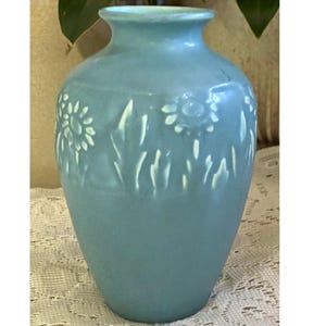 May include: A light blue ceramic vase with a floral design. The vase has a rounded body and a narrow neck. White flowers and leaves are embossed on the vase. The vase is approximately 8 inches tall.