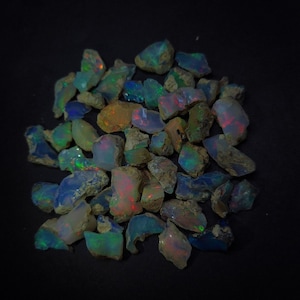 Op de afbeelding: Een verzameling ruwe, iriserende opaal edelstenen in verschillende tinten blauw, groen en rood. De opalen zijn onregelmatig van vorm en grootte, en sommige hebben een ruwe, natuurlijke textuur.