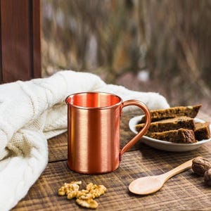 May include: A copper-colored mug with a handle sits on a wooden surface. Beside it are walnuts, a wooden spoon, and a plate of sliced bread. A white knitted blanket is draped nearby, creating a cozy, rustic scene.