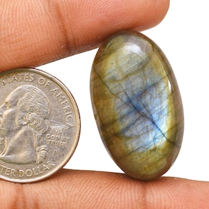 May include: A large, oval-shaped, green labradorite gemstone with blue and gold iridescent flashes. The gemstone is being held by a finger and is next to a US quarter.