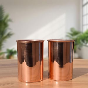 May include: Two copper-coloured cylindrical tumblers with a shiny finish. The tumblers have a simple, elegant design with a slightly flared rim. They are placed on a wooden surface, with a blurred background of greenery and a window.