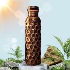 May include: A copper-coloured water bottle with a honeycomb pattern. The bottle has a screw-on cap and a cylindrical shape. The bottle is made of metal and has a shiny finish. The bottle is standing on a white pedestal with a natural background.