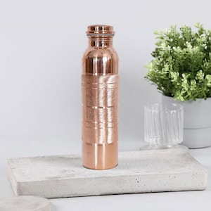 May include: A copper water bottle with a screw-on cap. The bottle has horizontal etched lines and a shiny finish. It is sitting on a concrete tray with a plant and glass in the background.