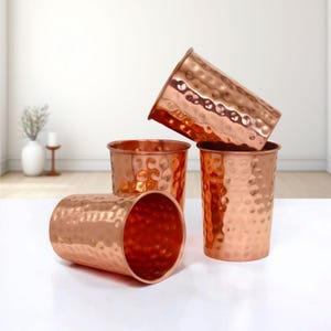 Peut inclure: Quatre gobelets de couleur cuivre avec une texture martelée. Les gobelets sont cylindriques avec un bord légèrement évasé. L'un des gobelets est couché sur le côté, tandis que les autres sont droits ou inclinés. Les gobelets sont disposés sur une surface blanche.