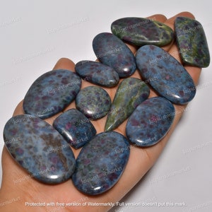 May include: A collection of polished gemstones in various shapes, including ovals and rectangles. The stones have a blue-grey base color with red and green inclusions. The gemstones are held in a hand.