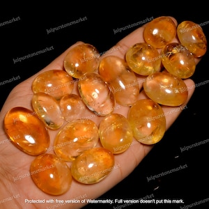 Może przedstawiać: Kolekcja 15 owalnych, prześwitujących, żółto-pomarańczowych kamieni szlachetnych z cytrynu. Kamienie są gładkie i wypolerowane, z lekkim połyskiem.