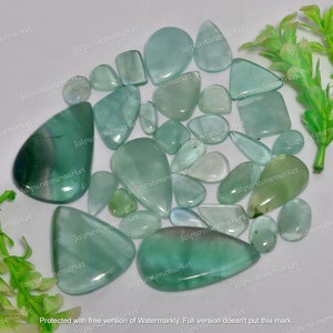 May include: A collection of polished gemstones in various shapes and sizes, showcasing shades of green and blue. The stones are arranged on a white surface, with some appearing translucent. The collection includes teardrop, oval, and rectangular shapes.