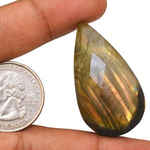 May include: A teardrop-shaped labradorite gemstone with a golden and green iridescent sheen. The gemstone is approximately the size of a US quarter.