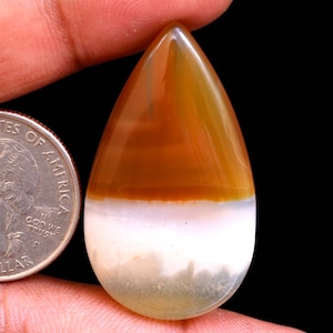 May include: A teardrop-shaped cabochon gemstone with a layered pattern of white, brown, and orange agate. The gemstone is approximately the size of a US quarter.