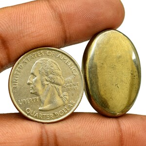 Pode incluir: Uma pedra de pirita ovalada de cor dourada ao lado de uma moeda de um quarto de dólar americano. A moeda apresenta o perfil de George Washington e as palavras "United States of America", "In God We Trust", "Liberty" e "Quarter Dollar".