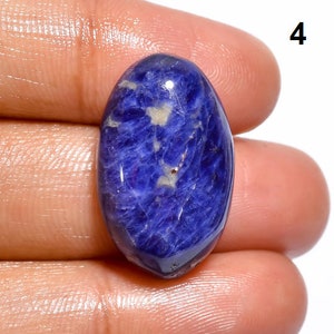 Op de afbeelding: Een close-up van een blauwe ovale cabochon edelsteen met witte en zilveren insluitsels. De edelsteen wordt in een hand gehouden.