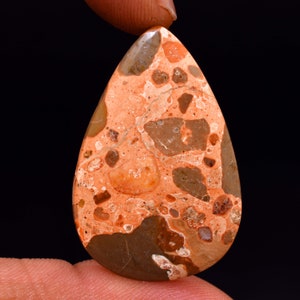 May include: A teardrop-shaped gemstone with a mottled pattern of orange, brown, and tan hues. The polished surface reveals a mix of textures and colors, creating a unique and natural appearance. The gemstone is held against a black background.