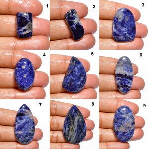 Op de afbeelding: Negen verschillende blauwe en witte cabochon edelstenen, elk met een uniek patroon en vorm. De edelstenen worden op een hand getoond, waarbij elke edelsteen van 1 tot en met 9 is genummerd.