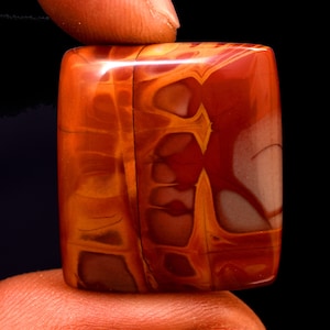 May include: A polished, rectangular gemstone with a rich color palette of deep red, orange, and cream. The stone's surface displays intricate patterns and veining, creating a unique and natural aesthetic. The gemstone is held between two fingers against a black background.