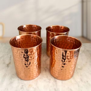 May include: Four copper-coloured drinking glasses with a hammered texture. The glasses are cylindrical with a slightly flared rim. The copper has a warm, reflective sheen, and the glasses are arranged on a light-coloured surface.